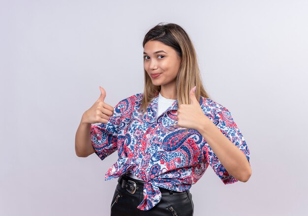 Una hermosa mujer joven con camisa estampada de paisley mostrando los pulgares hacia arriba mientras mira en una pared blanca