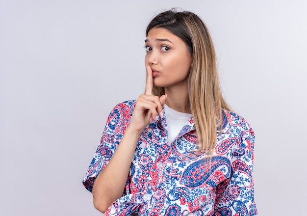 Una hermosa mujer joven con camisa estampada de paisley que muestra el gesto shh con el dedo mientras mira en una pared blanca