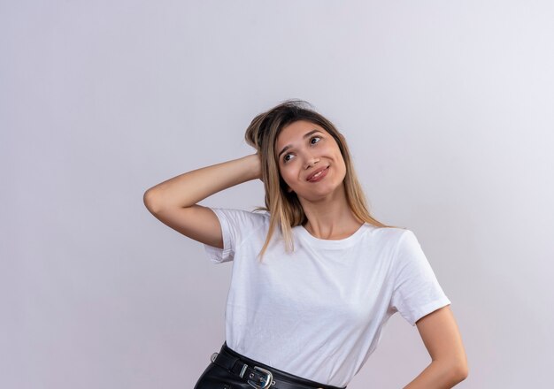 Una hermosa mujer joven en camiseta blanca pensando en algo agradable mientras mantiene la mano en la cabeza sobre una pared blanca