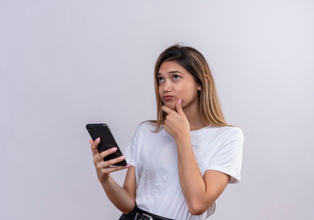 Una hermosa mujer joven en camiseta blanca pensando mientras sostiene el teléfono móvil en una pared blanca