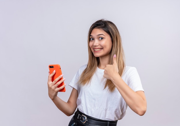 Una hermosa mujer joven en camiseta blanca que muestra los pulgares hacia arriba mientras sostiene el teléfono móvil en una pared blanca