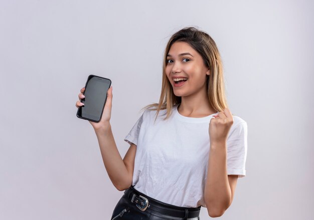 Una hermosa mujer joven en camiseta blanca sonriendo mientras muestra un espacio en blanco del teléfono móvil con el puño cerrado en una pared blanca