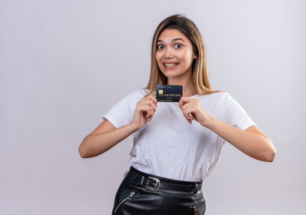 Una hermosa mujer joven en camiseta blanca sonriendo mientras muestra la tarjeta de crédito en una pared blanca