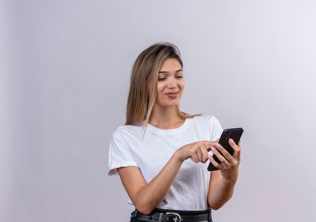 Una hermosa mujer joven en camiseta blanca sonriendo mientras toca la pantalla del teléfono móvil en una pared blanca