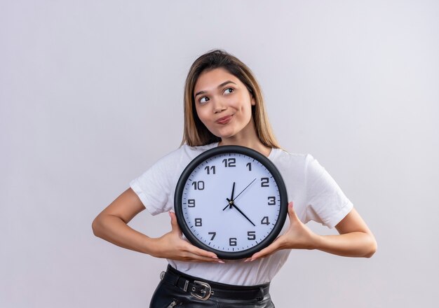 Una hermosa mujer joven en camiseta blanca sosteniendo el reloj de pared mientras mira de lado en una pared blanca