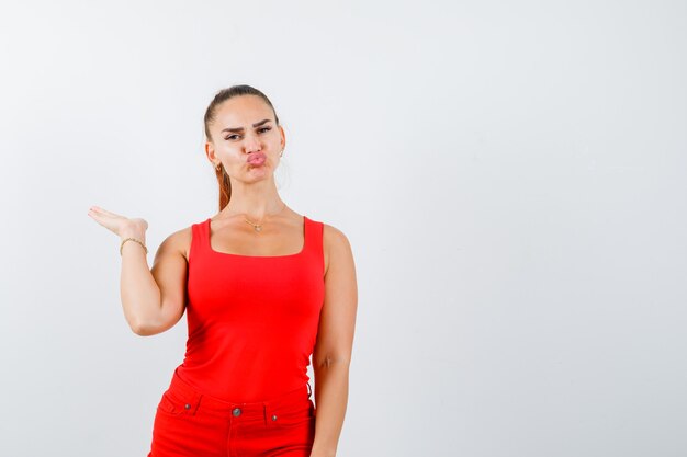 Hermosa mujer joven en camiseta sin mangas roja, pantalones pretendiendo sostener algo, haciendo pucheros con los labios y mirando confiado, vista frontal.