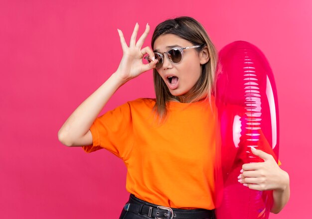 Una hermosa mujer joven con una camiseta naranja con gafas de sol que muestra un gesto de ok mientras sostiene un anillo inflable rosa en una pared rosa