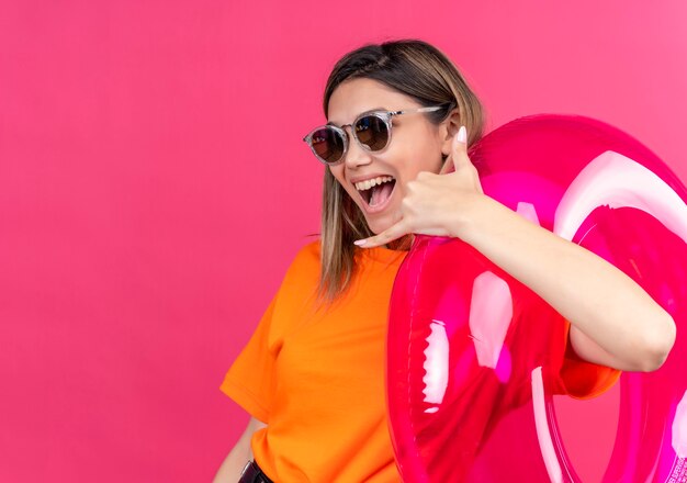 Una hermosa mujer joven con una camiseta naranja con gafas de sol sonriendo y mirando mostrando el gesto de llamarme mientras sostiene un anillo inflable rosa en una pared rosa