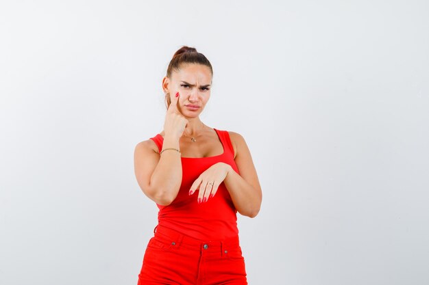 Hermosa mujer joven en camiseta roja, pantalones apuntando a su párpado y mirando molesto, vista frontal.