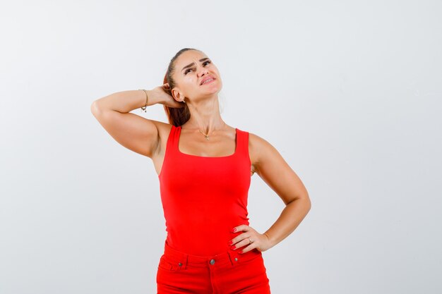 Hermosa mujer joven en camiseta roja, pantalones rascándose la cabeza mientras mira hacia arriba y mira ansiosa, vista frontal.