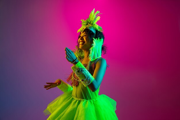Hermosa mujer joven en carnaval, elegante disfraz de mascarada con plumas bailando en la pared degradada en neón