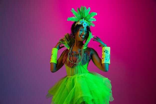 Hermosa mujer joven en carnaval, elegante disfraz de mascarada con plumas bailando sobre fondo degradado en neón.
