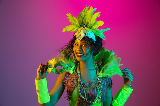 Hermosa mujer joven en carnaval, elegante disfraz de mascarada con plumas bailando sobre fondo degradado en neón.