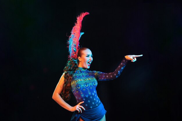 Hermosa mujer joven en carnaval, elegante disfraz de mascarada con plumas en pared negra en luz de neón