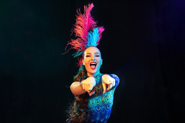 Hermosa mujer joven en carnaval, elegante disfraz de mascarada con plumas en pared negra en neón