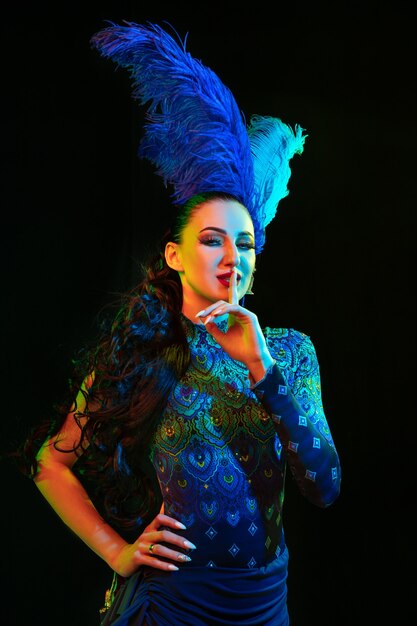 Hermosa mujer joven en carnaval, elegante disfraz de mascarada con plumas sobre fondo negro en luz de neón.