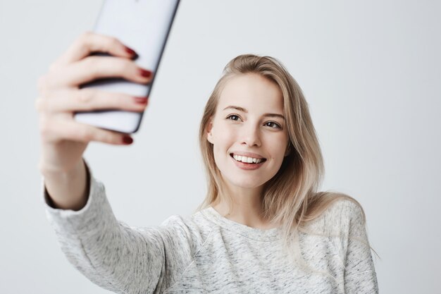 Hermosa mujer joven caucásica con largo cabello rubio teñido y atractivos ojos oscuros con teléfono móvil, posando para selfie