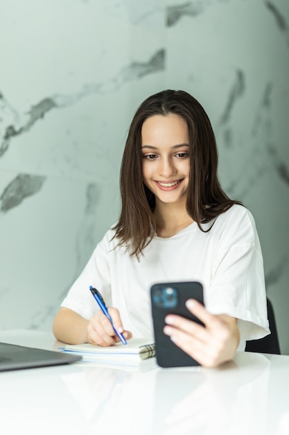 Foto gratuita hermosa mujer joven concentrada escribiendo mensajes de texto en el teléfono móvil mientras calcula facturas en la cocina, sentado a la mesa con computadora portátil, papeles y calculadora