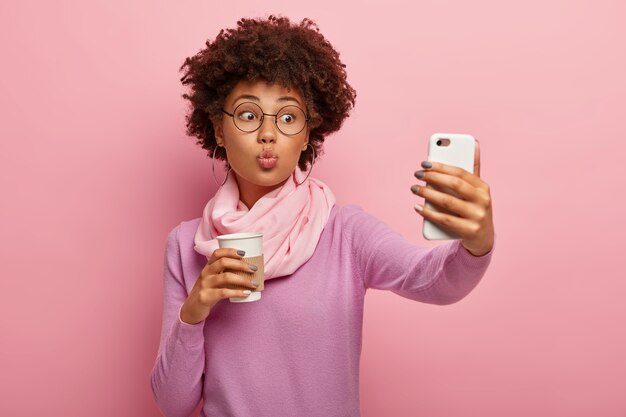 Hermosa mujer joven con corte de pelo afro, mantiene los labios doblados, sopla beso en la cámara del teléfono inteligente, toma retrato selfie