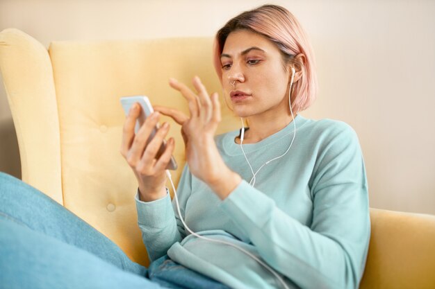 Hermosa mujer joven elegante con cabello rosado sentado cómodamente en un sillón con teléfono celular