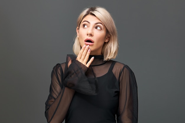 Hermosa mujer joven con un elegante corte de pelo y una elegante blusa negra después de haber sorprendido la expresión facial de sorpresa, mirando hacia arriba con la boca abierta, sin palabras, no puedo ocultar el asombro y la conmoción