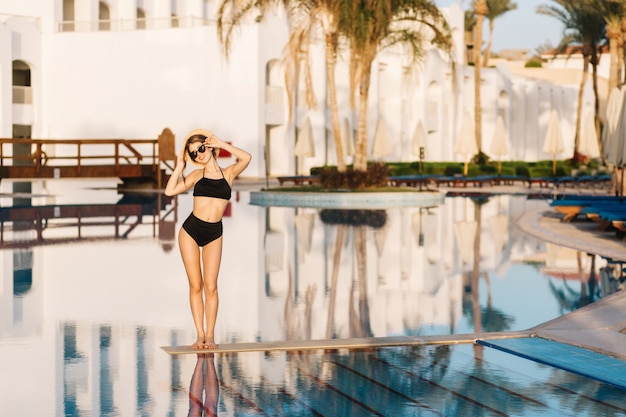 Hermosa mujer joven con elegante traje de baño negro, bikini, chica de vacaciones, vacaciones, resort, en un bonito hotel. El uso de elegantes gafas de sol, sombrero de paja en la mano.