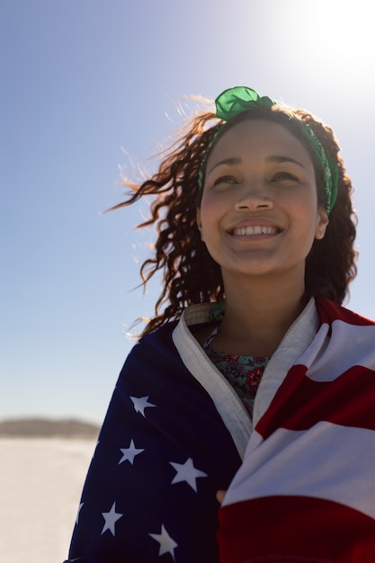 Hermosa mujer joven envuelta en la bandera americana en la playa bajo el sol