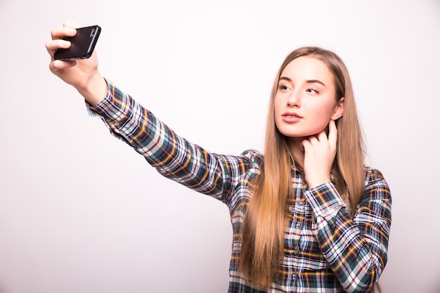 Hermosa mujer joven está haciendo foto selfie con smartphone aislado en pared blanca