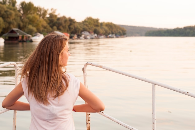 Hermosa mujer joven está mirando a la puesta de sol sobre el río Atractiva mujer disfruta de verano y cálida luz del sol. Ella está admirando la vista hermosa y disfrutando de la puesta del sol. El viento le soplaba el pelo.