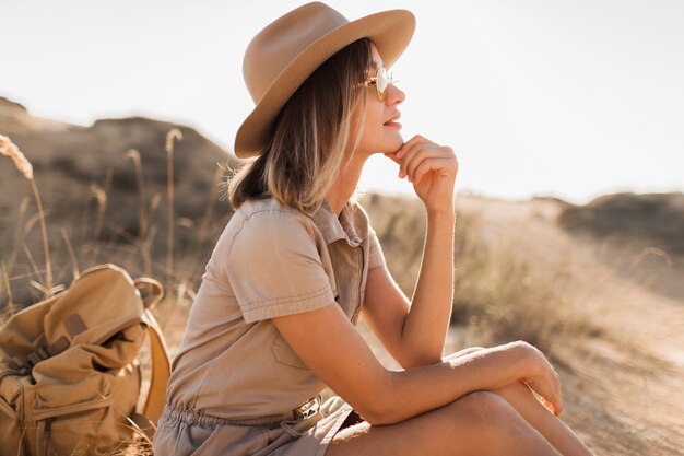 Hermosa mujer joven con estilo en vestido caqui en la arena del desierto viajando en África en safari con sombrero y mochila