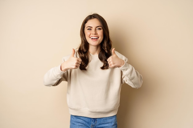 Hermosa mujer joven, estudiante mostrando los pulgares para arriba en aprobación, recomendando la tienda, de pie sobre fondo beige