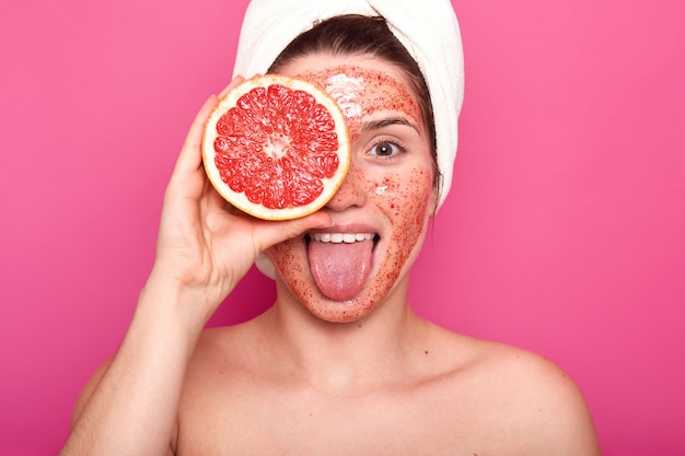 Hermosa mujer joven con exfoliante brillante en la cara sostiene la mitad de la toronja de violación en una mano, saca la lengua, con una toalla blanca en la cabeza, se ve fresca y encantada. Concepto de cuidado de la piel.