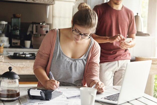 Hermosa mujer joven con gafas mirando seriamente escribiendo con bolígrafo mientras administra impuestos y calcula facturas, tratando de reducir los gastos domésticos para ahorrar dinero y permitirse hacer una gran compra