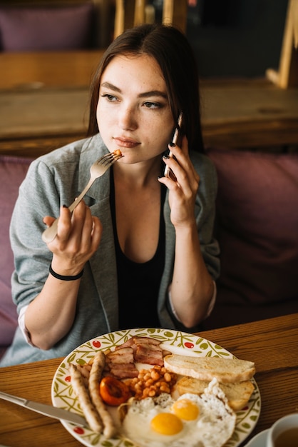 Hermosa mujer joven hablando por teléfono móvil comiendo alimentos con tenedor