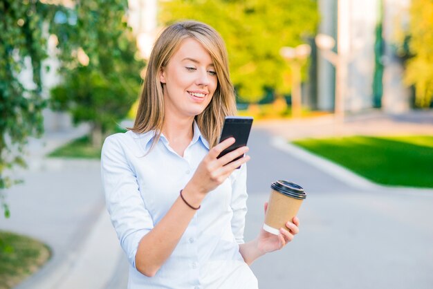 Hermosa mujer joven hermosa con el pelo rubio de mensajería en el teléfono inteligente en el fondo de la calle de la ciudad. Chica bonita que tiene conversación telefónica inteligente en llamarada del sol.