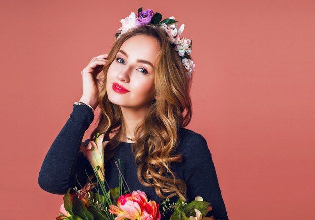 Hermosa mujer joven con largo cabello rubio ondulado en guirnalda de flores de primavera posando con ramo de flores sobre fondo rosa.