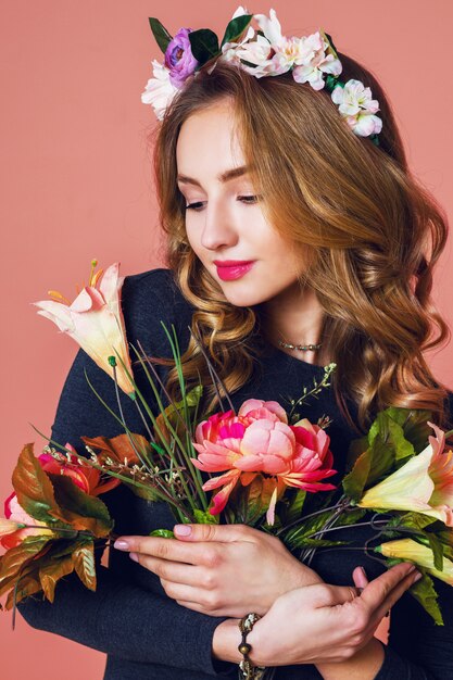 Hermosa mujer joven con largo cabello rubio ondulado en guirnalda de flores de primavera posando con ramo de flores sobre fondo rosa.