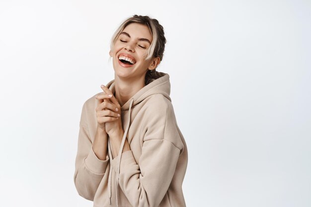 Hermosa mujer joven con maquillaje natural riendo y sonriendo despreocupada expresión de cara feliz de pie en sudadera con capucha sobre fondo blanco.