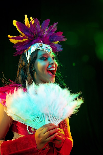 Hermosa mujer joven en máscara de carnaval y elegante traje de mascarada con abanico de plumas en luces de colores y resplandor sobre fondo negro.