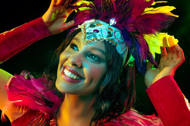 Hermosa mujer joven en máscara de carnaval y elegante traje de mascarada con plumas en luces de colores y resplandor sobre fondo negro.