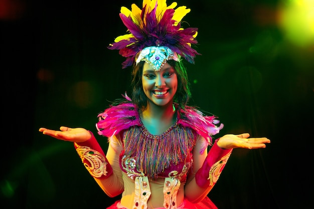 Hermosa mujer joven en máscara de carnaval y elegante traje de mascarada con plumas en luces de colores y resplandor sobre fondo negro.