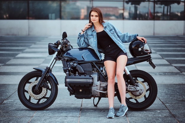 Hermosa mujer joven en minishorts y chaqueta de mezclilla está posando en la bicicleta mientras sostiene el casco.