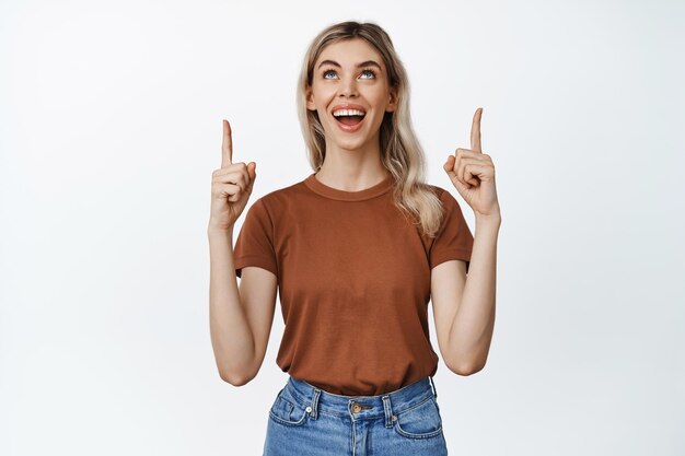 Hermosa mujer joven mirando algo con emoción apuntando con el dedo hacia arriba y mirando hacia arriba en el anuncio de la tienda del logotipo de pie contra el fondo blanco