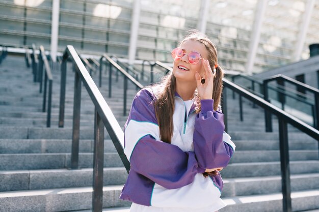 Hermosa mujer joven de moda en ropa elegante con gafas de sol rosas y sonriendo