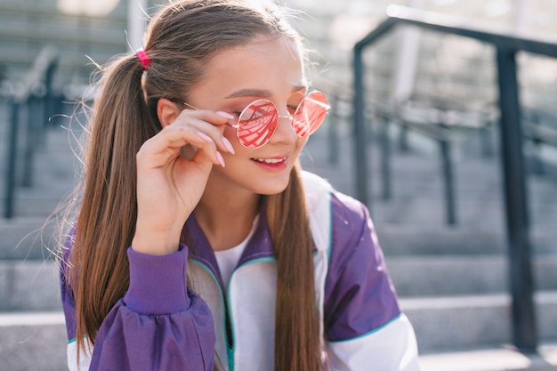 Hermosa mujer joven de moda en ropa elegante con gafas de sol rosas y sonriendo