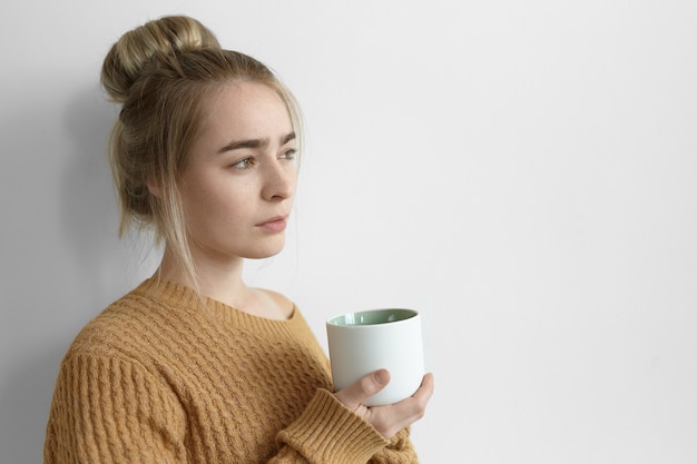 Hermosa mujer joven con moño sosteniendo una taza grande de té, mirando hacia delante con expresión facial pensativa. Linda mujer en suéter acogedor tomando café