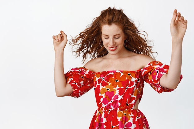 Hermosa mujer joven con peinado de jengibre rizado vestido floral de verano bailando y sonriendo despreocupada mirando hacia abajo feliz de pie sobre fondo blanco Copiar espacio