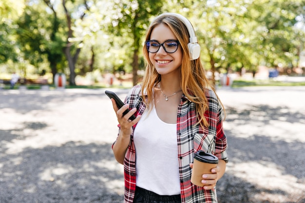 Hermosa mujer joven de pie al aire libre con una taza de café. Retrato de niña con estilo de buen humor en auriculares.