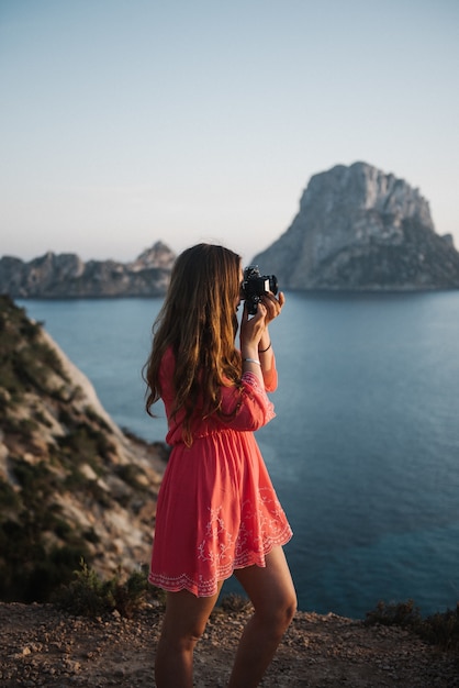 Hermosa mujer joven de pie junto al mar tomando una foto con una cámara