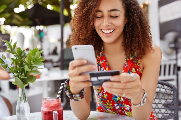 Hermosa mujer joven de piel oscura con expresión alegre, tiene teléfono inteligente y tarjeta de crédito, bancos en línea o hace compras mientras se sienta contra el interior del café.